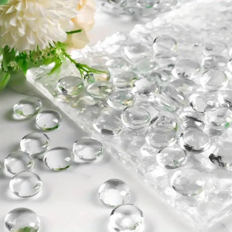 Clear glass pebbles from STONED scattered on a white surface with a white flower in the background SGI Online